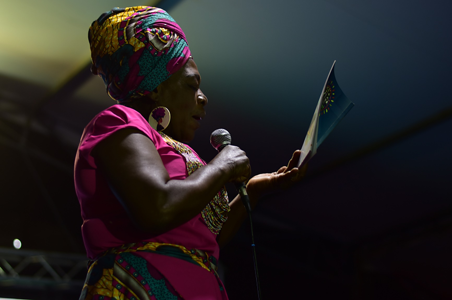 Mujer afrodescendiente con turbante de colores, aretes grandes y vestido fucsia habla al micrófono en un escenario de Afrotex mientras lee un folleto