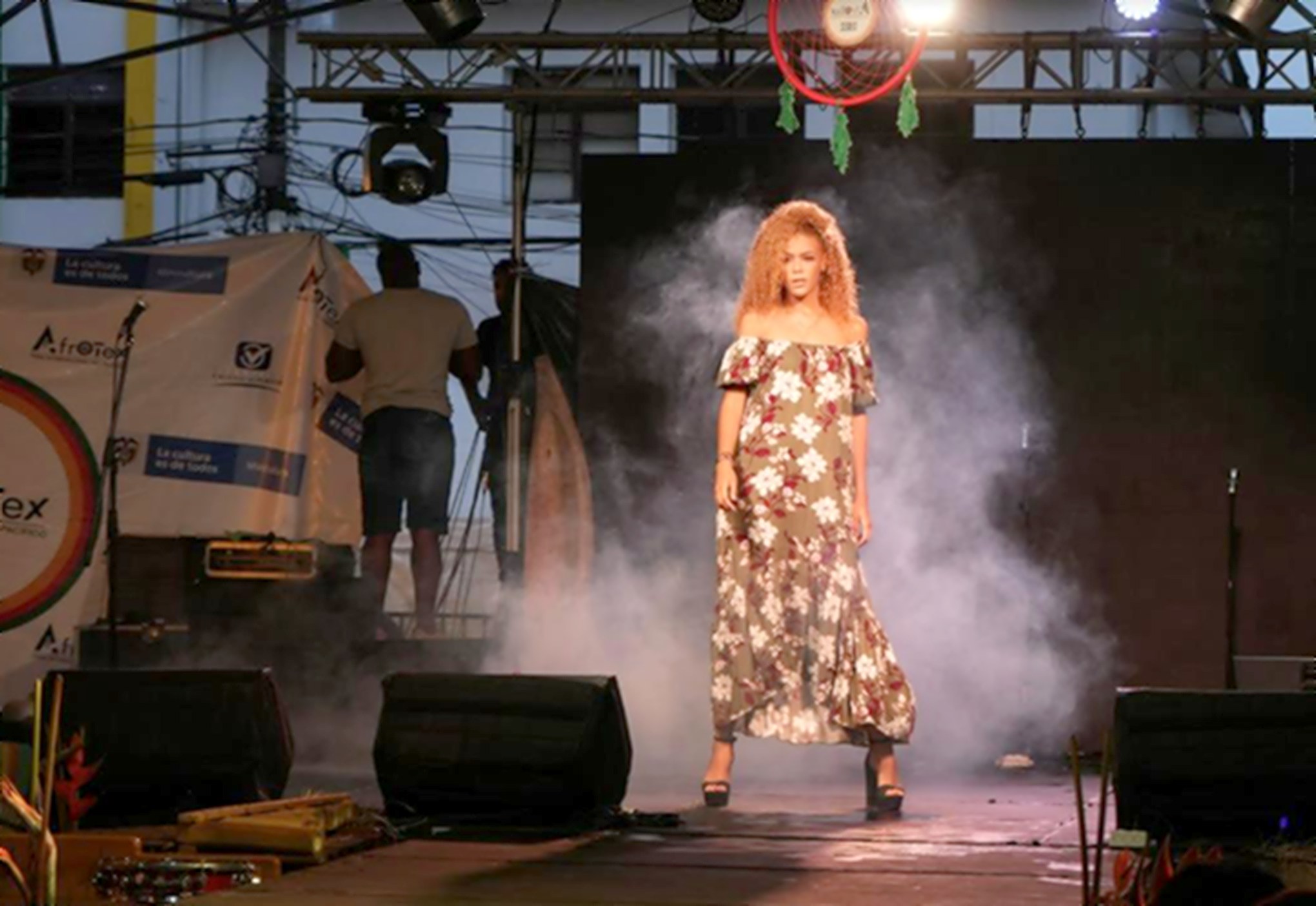 Modelo de cabello rizado con vestido largo de flores desfila entre humo y luces en la pasarela de Afrotex