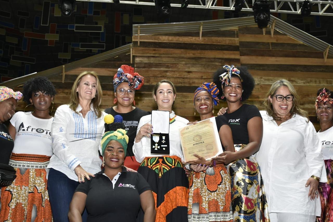 Mujeres de Afrotex con camisetas de la feria, turbantes y faldas estampadas, sosteniendo una medalla y un diploma de reconocimiento en un escenario