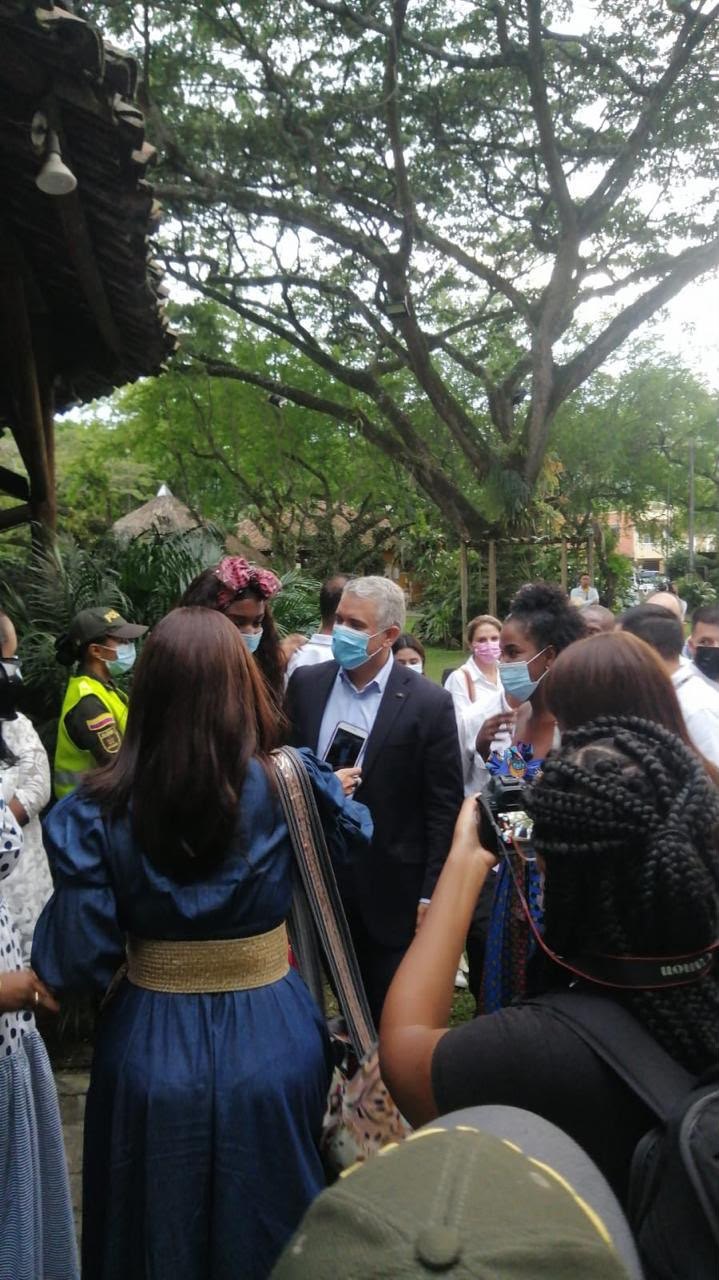 Grupo de personas con tapabocas al aire libre bajo un gran árbol, conversando durante una visita; una joven con trenzas toma fotos